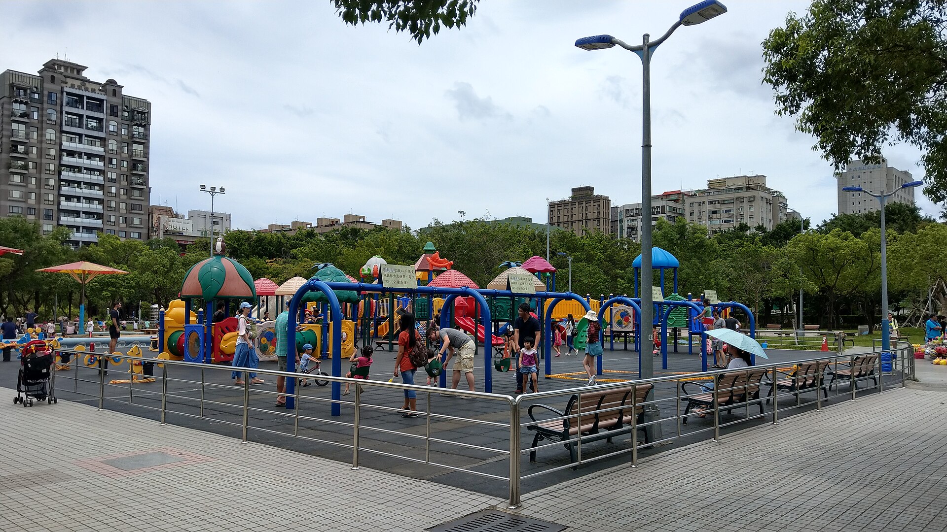 台北大安森林公園的兒童遊戲場,彩色溜滑梯與遊具前有幾位家長和小孩,背景是公寓大樓。