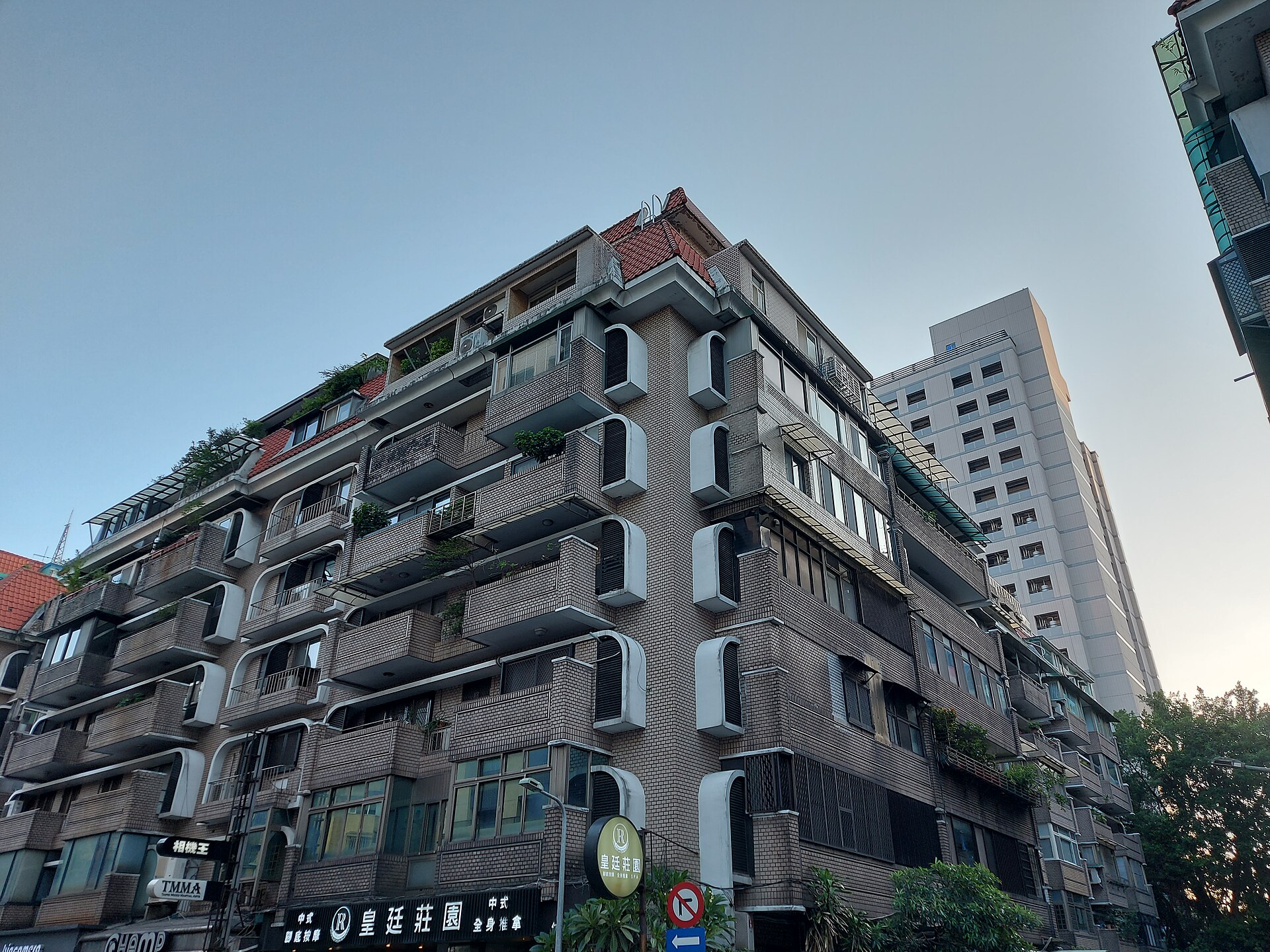 從街角仰拍台北一棟紅瓦頂、磚色外牆的老公寓,每層陽台外掛著冷氣,後方是一棟白色高樓。